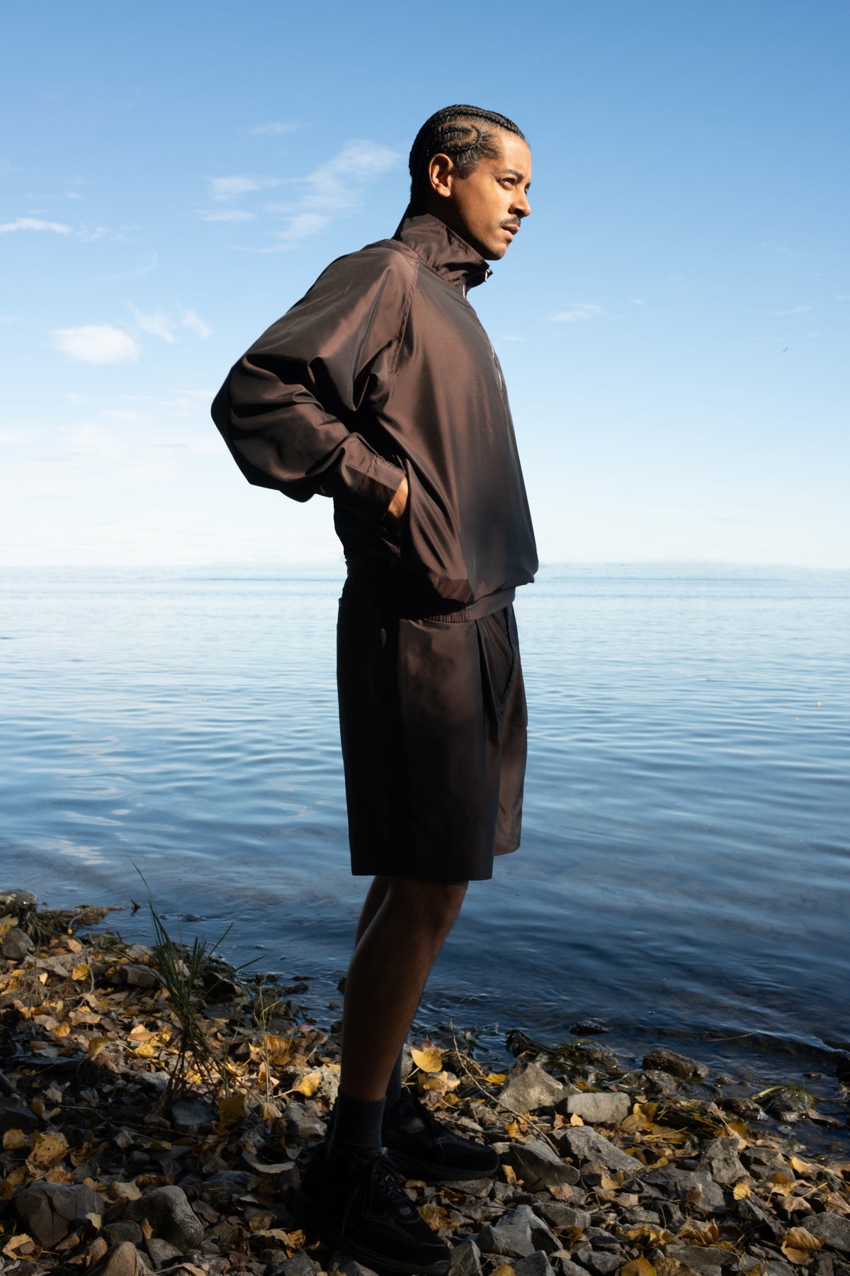 Pierre FW26 campaign, Nylon Silk Quarter Zip Jacket in Brown, Nylon Silk shorts in brown, standing by the water